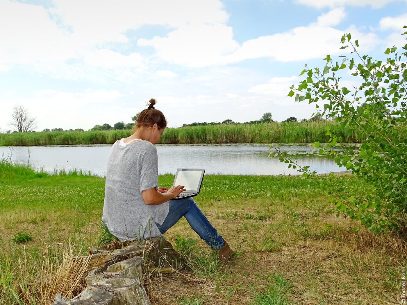 Frau sitzt an einem See und arbeitet am Laptop