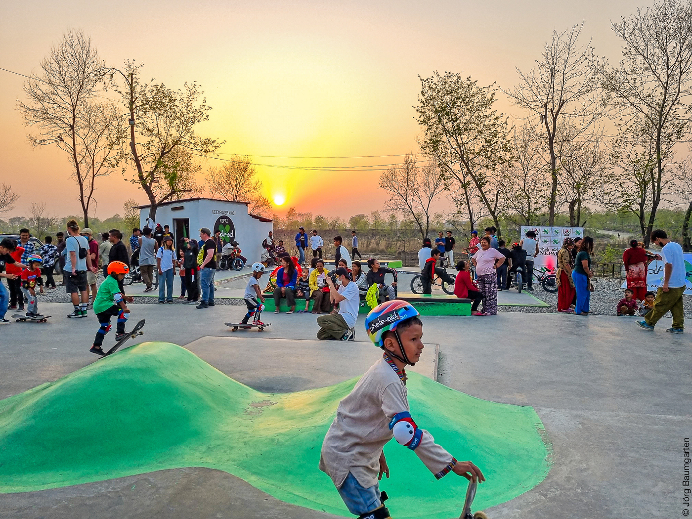 Das Foto zeigt einen Jungen mit einem bunten Schutzhelm auf einem Skatepark, sowie Publikum