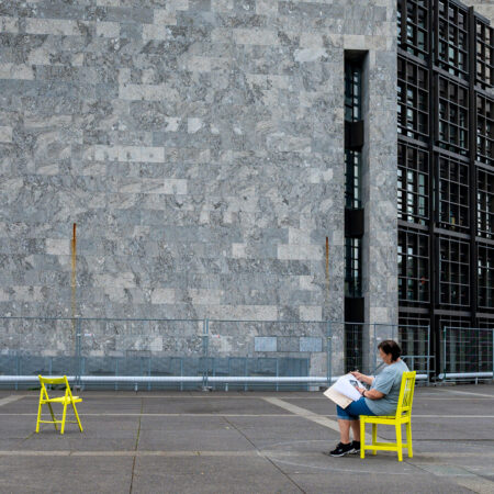 Frau sitzt auf einem gelben Stuhl vor der steinernen Fassade des Arne Jacobsen Rathauses in Mainz.