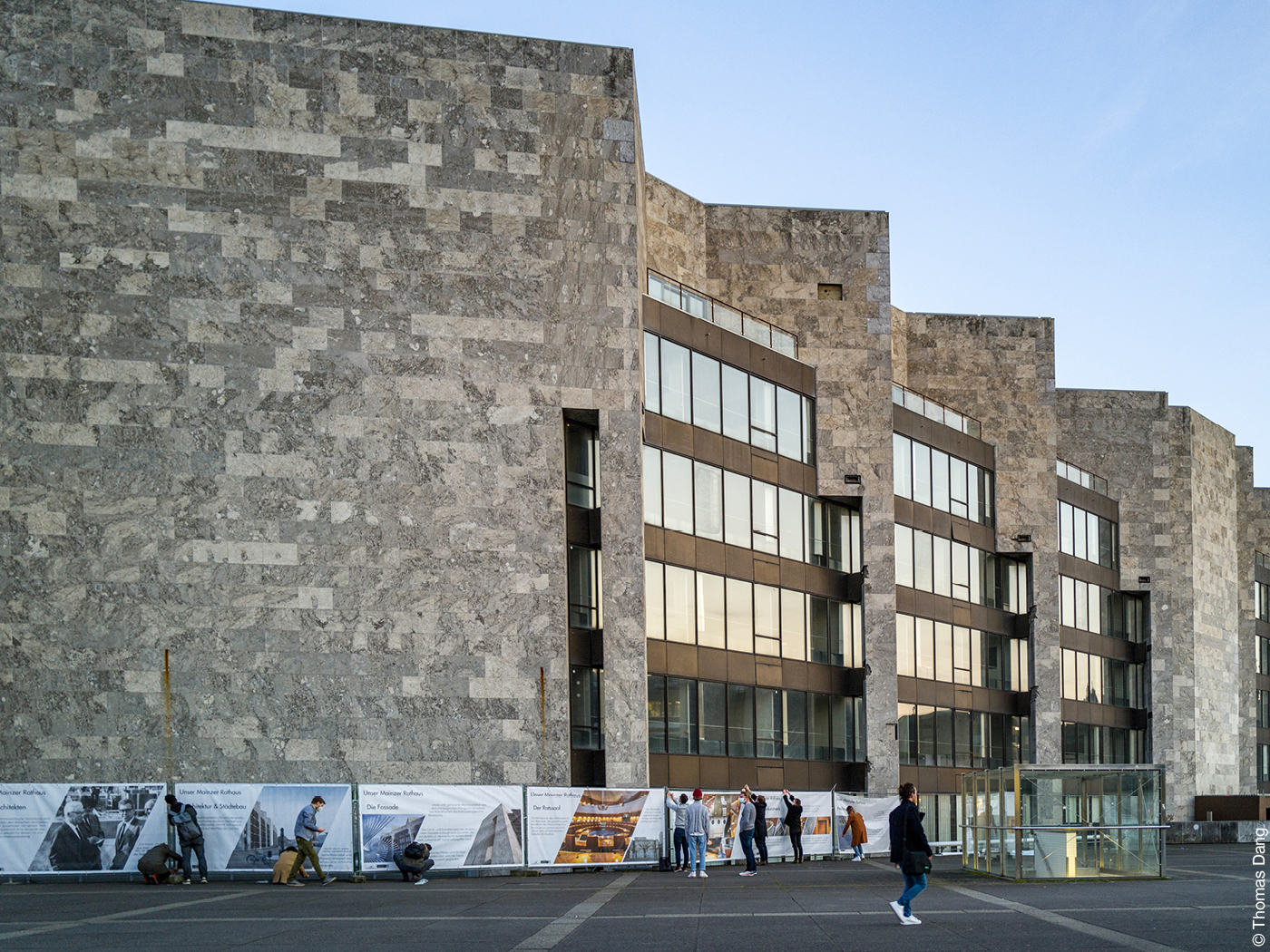 Steinfassade des Arne Jacobsen Rathauses in Mainz, davor Ausstellungsplakate über das Gebäude und Passanten.