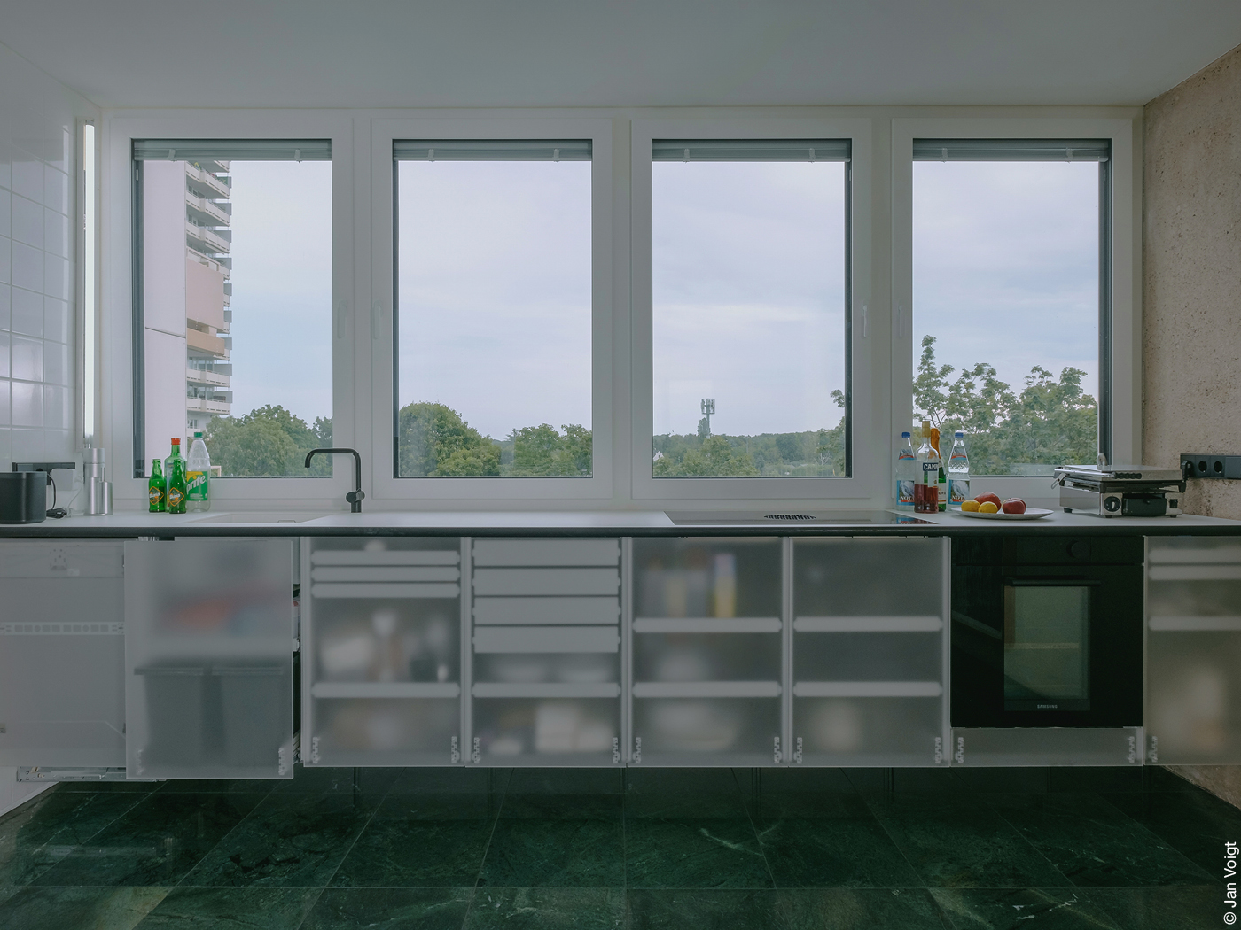 Moderne Küchenzeile mit Milchglas-Küchenschränken und einer breiten Fensterfront, durch die man auf Baumwipfel blickt.