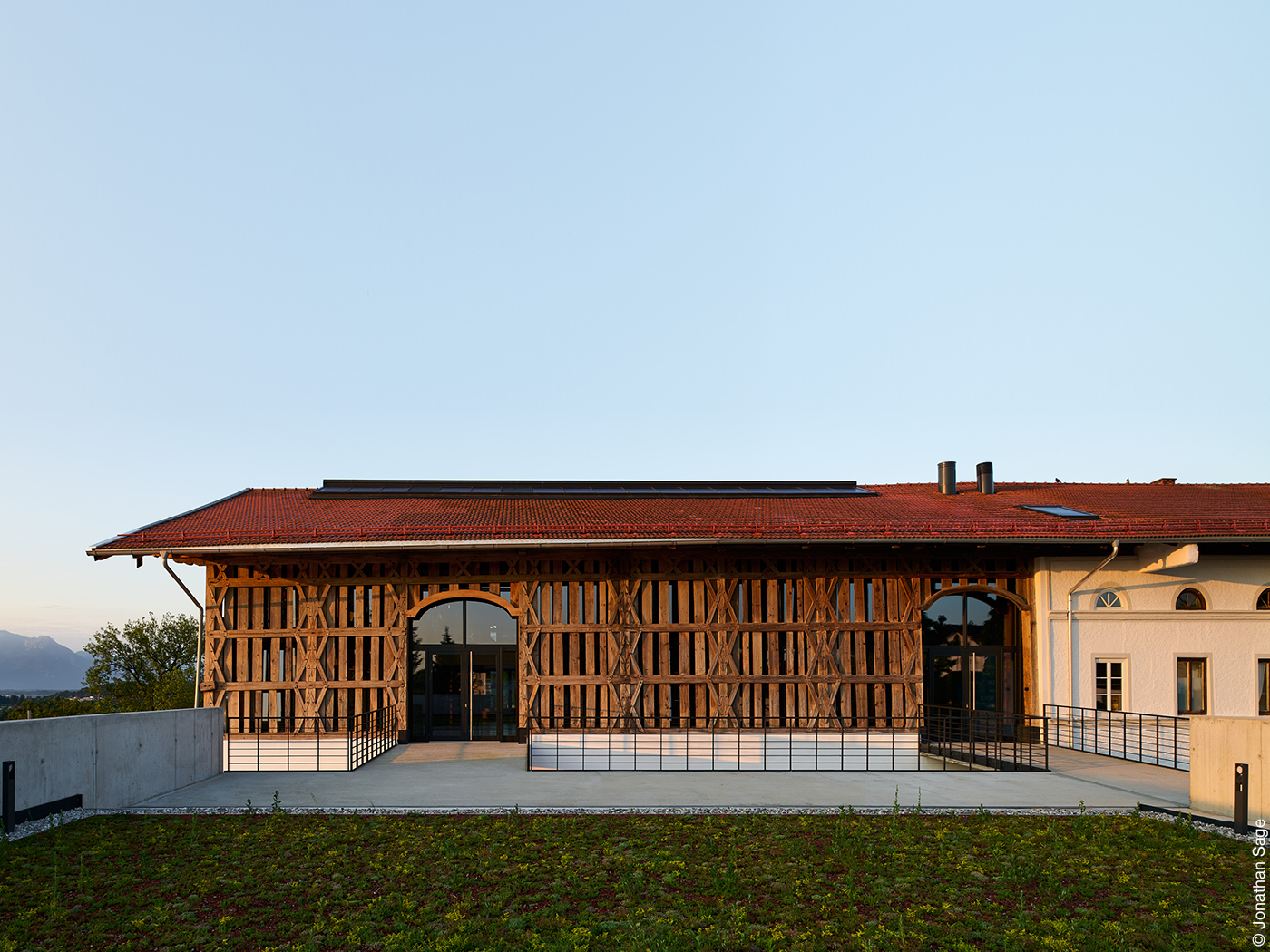 Flaches Haus mit kunstvoller Holzfassade, davor Rasen, dahinter Berge