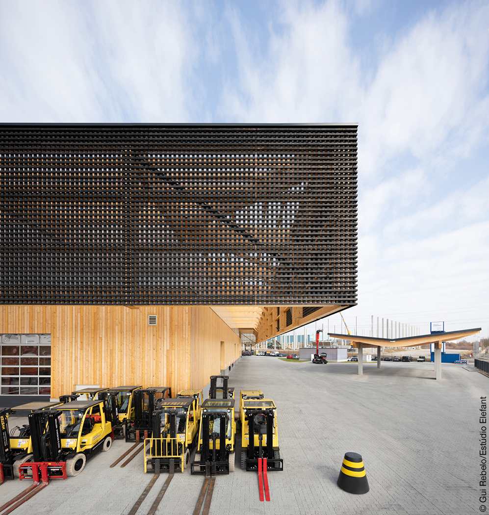 Industriehalle mit weit auskragendem Vordach aus schwarzem Holz mit Gittermuster, davor stehen mehrere Gabelstapler