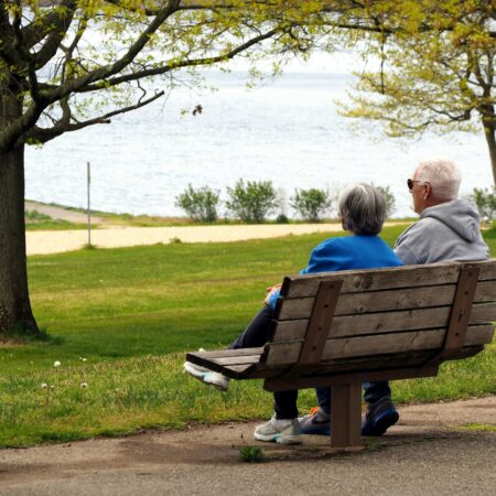 Ein älteres Paar sitzt in einem Park auf einer Bank und blickt auf einen See.