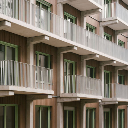 Wohngebäude mit Holzfassade, quadratischen Fenstern mit grünen Fensterrahmen und weißen Balkons